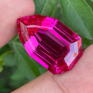 Può includere: Una gemma rosa intenso e vibrante con una forma sfaccettata a scudo. La pietra presenta tagli geometrici netti che creano riflessi interni e una finitura lucida, mostrando una tonalità magenta brillante e satura in tutta la struttura.