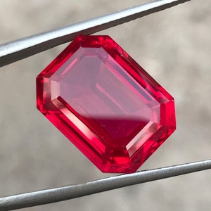 May include: A vibrant red, rectangular-cut gemstone, held by silver tweezers. The gem's facets catch the light, creating a sparkling effect. The tweezers are in focus, emphasizing the gemstone's clarity and the precision of the cut.