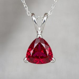 May include: A silver pendant necklace featuring a triangular, faceted, deep red gemstone. The pendant is suspended from a delicate silver chain. The gemstone is set in a silver bezel setting, creating a striking contrast.