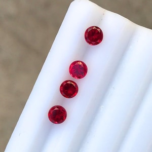 May include: Four round, faceted, deep red gemstones are arranged diagonally on a white surface. The gems have a brilliant cut, reflecting light. The gemstones are likely rubies, suitable for jewelry making or collecting.