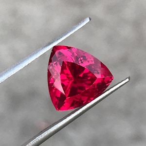 Puede incluir: Una gema de color rojo intenso y vibrante, tallada en forma triangular con múltiples facetas que reflejan la luz. La gema está sujeta por unas pinzas de metal plateado, lo que resalta su color brillante y su corte geométrico preciso.