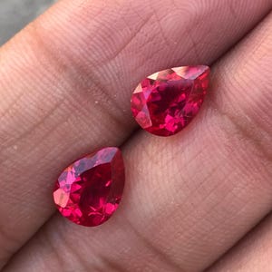 May include: Two faceted, pear-shaped, bright red gemstones. The gems are displayed against a skin-toned background, showcasing their vibrant color and cut. The gems are likely rubies or similar precious stones, perfect for jewelry.