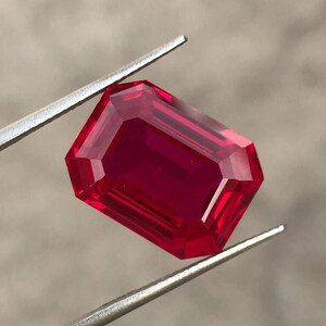 May include: A faceted, rectangular ruby gemstone held by silver tweezers. The ruby has a deep red color and a brilliant, reflective surface. The gemstone's cut features multiple facets, enhancing its sparkle and clarity. The tweezers provide a contrasting metallic element.