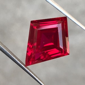 May include: A vivid red, kite-shaped gemstone, held by silver tweezers. The cut of the gem reveals multiple facets, enhancing its brilliance. The gemstone is set against a neutral background. The gem's shape is reminiscent of a kite.