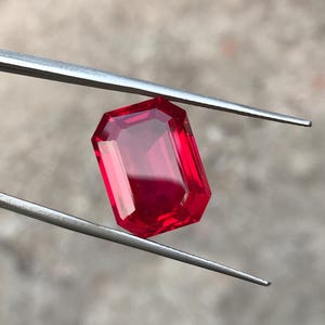 May include: A vibrant, transparent red gemstone with an emerald-cut shape, featuring sharp, clean facets that reflect light. The rectangular stone is held securely by a pair of thin, silver-toned metal tweezers, showcasing its deep crimson color.