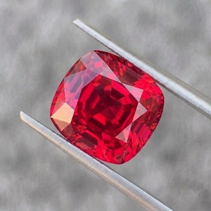 May include: A vibrant, cushion-cut red gemstone held by silver-toned metal tweezers. The stone features a faceted surface that reflects light, creating deep crimson and bright scarlet highlights across its square-like, rounded shape.
