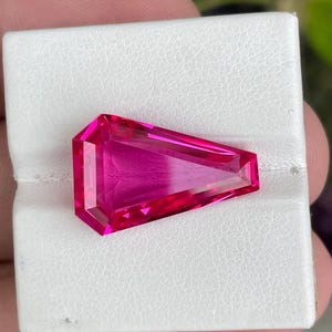 May include: A vibrant, hot pink gemstone cut into a shield-like, elongated pentagonal shape. The stone features precise, multi-faceted surfaces that reflect light, creating a brilliant, transparent appearance with deep magenta and bright pink hues.