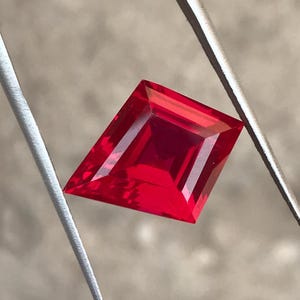 May include: A vibrant, faceted red gemstone with a unique kite-like shape. The gem is held by a silver tool, showcasing its brilliant cut and intense color. The gemstone's facets reflect light, creating a dazzling display.