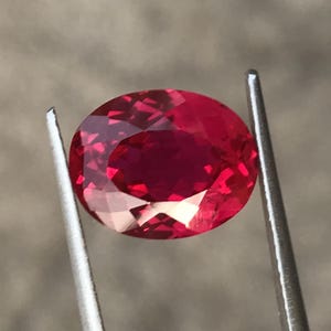 May include: A vibrant, oval-cut gemstone with a deep, saturated red color and multifaceted surface that reflects light. The stone is held securely between the two thin, silver-toned metal prongs of a pair of tweezers, showcasing its clarity.
