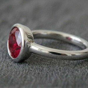 May include: A silver ring with a large, oval-cut, deep red gemstone. The ring's band is smooth and polished, and the gemstone is set in a bezel setting. The ring is displayed on a gray surface.