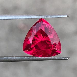 May include: A vibrant, deep red gemstone cut into a triangular shape with multiple facets that reflect light. The stone is held securely between the two metal tips of a pair of tweezers, showcasing its brilliant color and precise geometric cut.