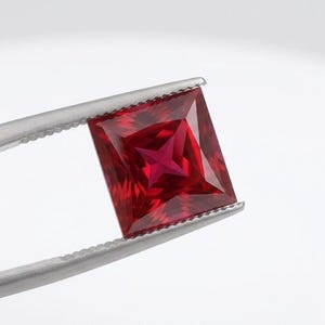 May include: A square-cut, vibrant red gemstone held by silver-toned metal tweezers. The stone features a faceted surface with a distinct four-pointed star pattern reflecting light from its center, showcasing deep crimson and bright red hues.