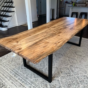 May include: A large wooden dining table with a live edge top and black metal legs. The table is sitting on a patterned rug.