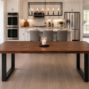 May include: A rectangular dining table featuring a thick, dark brown wooden top with a natural grain pattern, supported by two sturdy, black metal U-shaped legs. A single white pillar candle in a clear glass holder sits centred on the table.