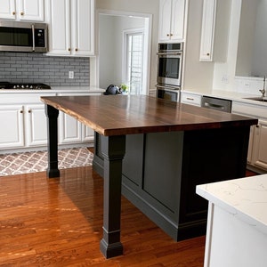May include: A dark wood kitchen island with a black base and a light wood countertop. The island has four legs and a raised edge around the countertop. The island is in a kitchen with white cabinets and hardwood floors.