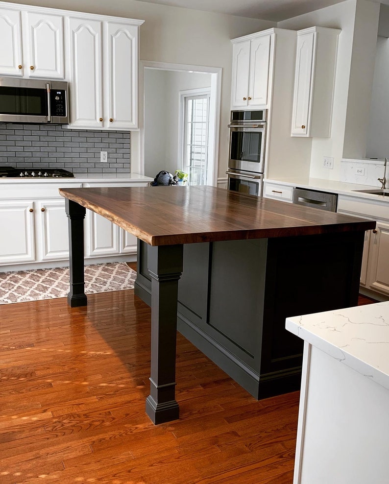 May include: A dark wood kitchen island with a black base and a light wood countertop. The island has four legs and a raised edge around the countertop. The island is in a kitchen with white cabinets and hardwood floors.