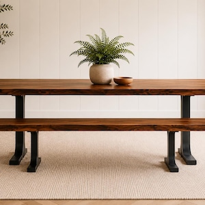 May include: A rectangular wooden dining table with a natural live edge and a matching bench, both featuring black metal legs. A potted green fern in a light grey ceramic planter and a small wooden bowl sit on the table surface.