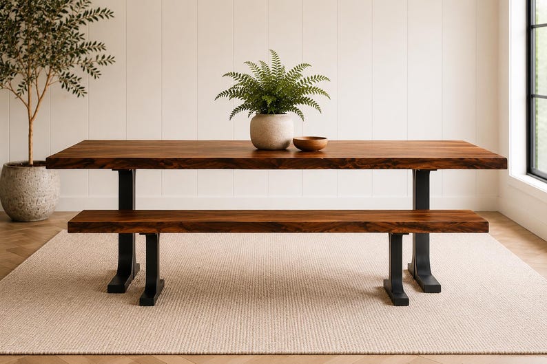 May include: A rectangular wooden dining table with a natural live edge and a matching bench, both featuring black metal legs. A potted green fern in a light grey ceramic planter and a small wooden bowl sit on the table surface.