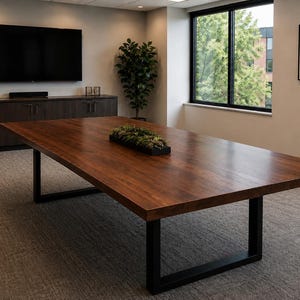 May include: A large rectangular conference table with a dark brown wood grain surface and black metal U-shaped legs. A long, narrow black rectangular planter filled with green artificial succulents sits in the centre of the table surface.