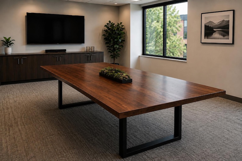 May include: A large rectangular conference table with a dark brown wood grain surface and black metal U-shaped legs. A long, narrow black rectangular planter filled with green artificial succulents sits in the centre of the table surface.