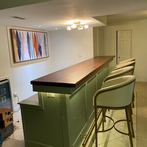May include: A green bar with a wood countertop and gold bar stools. The bar has a built-in shelf and a light under the countertop. The bar is in a basement with a white ceiling and a painting on the wall.