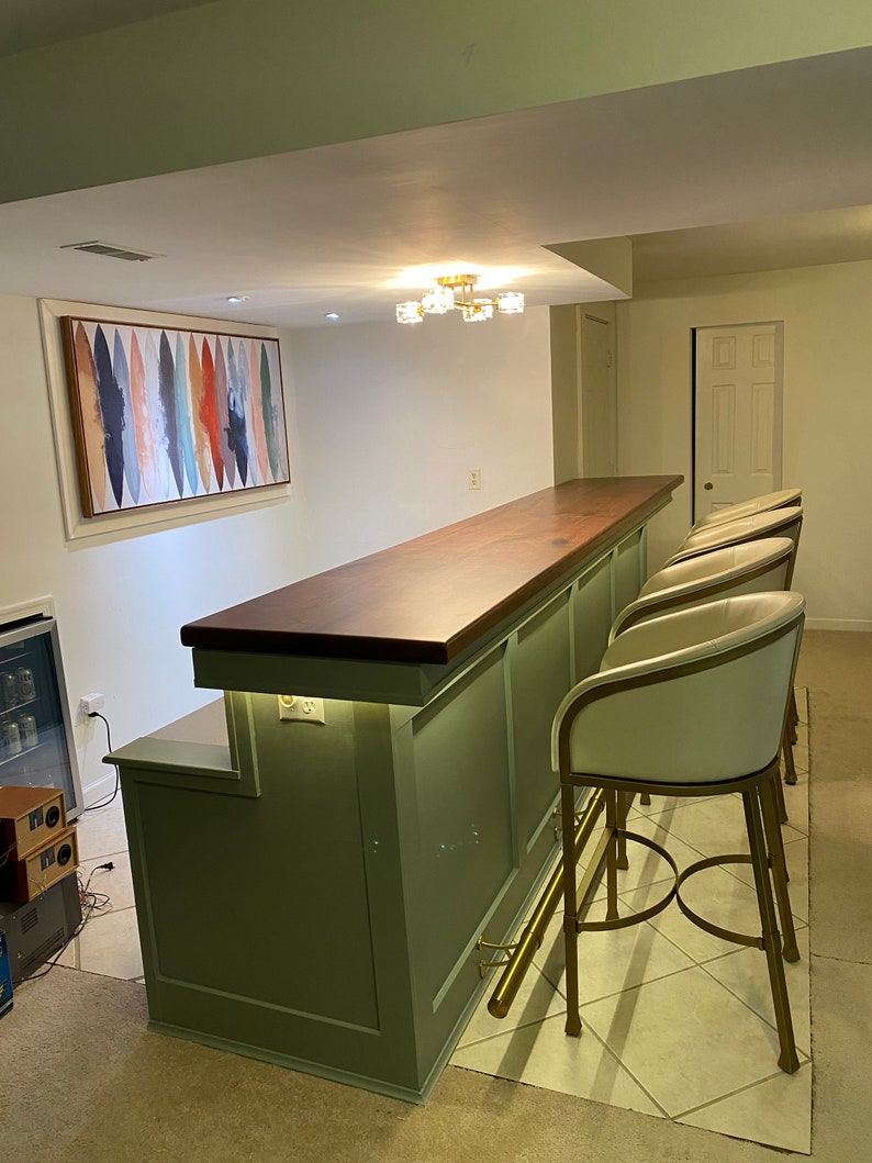 May include: A green bar with a wood countertop and gold bar stools. The bar has a built-in shelf and a light under the countertop. The bar is in a basement with a white ceiling and a painting on the wall.