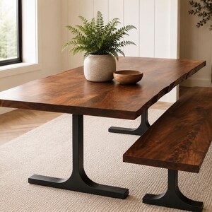 May include: A rectangular dining table with a natural live edge wood top and two black metal T-shaped pedestal bases, accompanied by a matching wooden bench. A light grey ceramic vase with a green fern and a small wooden bowl sit on the table.
