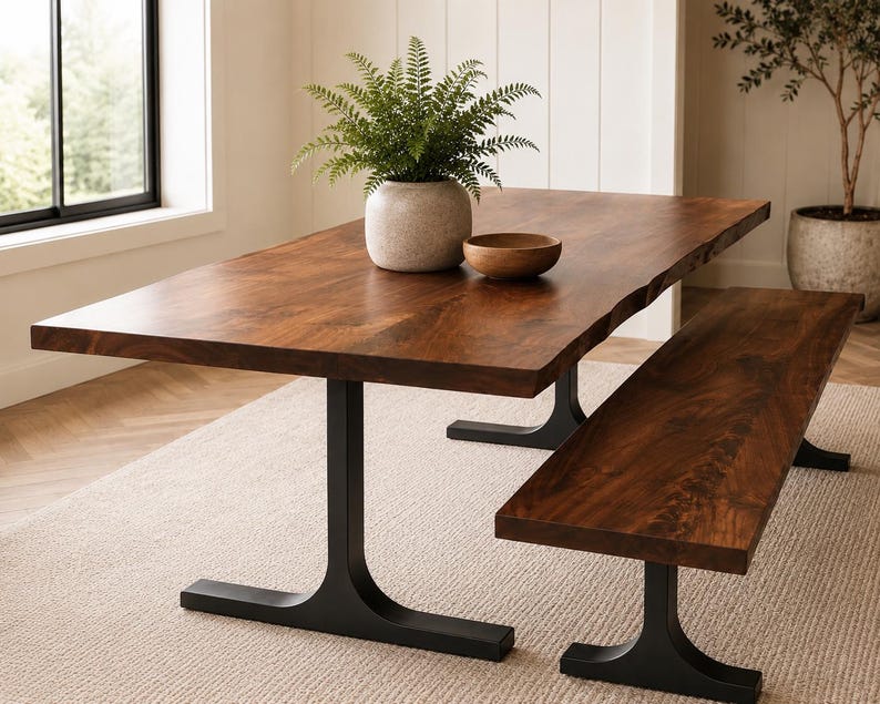 May include: A rectangular dining table with a natural live edge wood top and two black metal T-shaped pedestal bases, accompanied by a matching wooden bench. A light grey ceramic vase with a green fern and a small wooden bowl sit on the table.