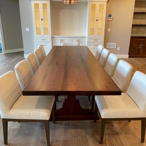 May include: A large, rectangular, wooden dining table with a dark brown finish. The table has a pedestal base and is surrounded by white upholstered chairs. The table is in a dining room with white cabinets and a light brown hardwood floor.