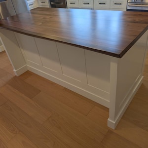May include: A white kitchen island with a dark wood countertop. The island has a simple design with four panels on the front and a base with a decorative molding.