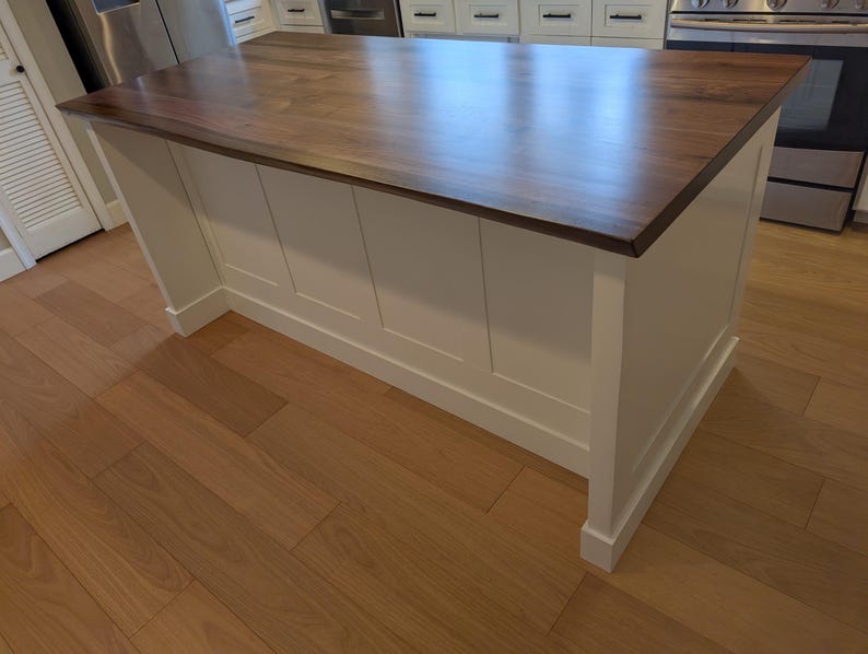 May include: A white kitchen island with a dark wood countertop. The island has a simple design with four panels on the front and a base with a decorative molding.
