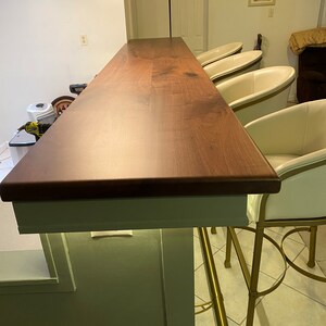 May include: A dark wood bar counter with a white edge and a light green wall behind it. Four white chairs with gold metal frames are positioned around the counter.