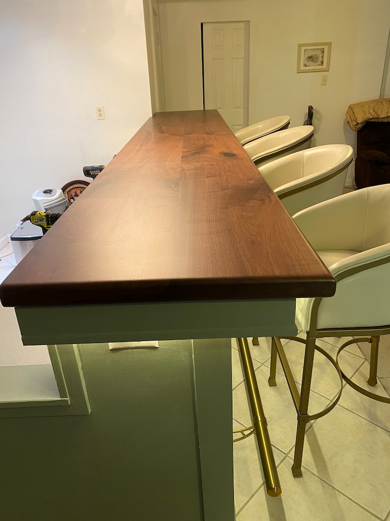 May include: A dark wood bar counter with a white edge and a light green wall behind it. Four white chairs with gold metal frames are positioned around the counter.