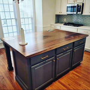 May include: A kitchen island with a dark gray base and a wood countertop. The island has three drawers and two doors with gold handles. The island is in a kitchen with hardwood floors.