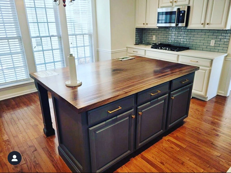 May include: A kitchen island with a dark gray base and a wood countertop. The island has three drawers and two doors with gold handles. The island is in a kitchen with hardwood floors.