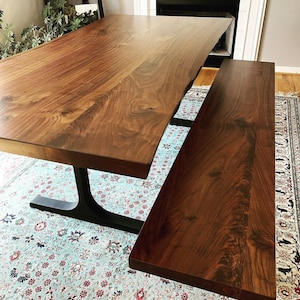 May include: A large rectangular dining table and a matching bench made of dark brown wood with a visible natural grain pattern. The table features a live edge side and is supported by a black metal base with a curved, pedestal-style design.