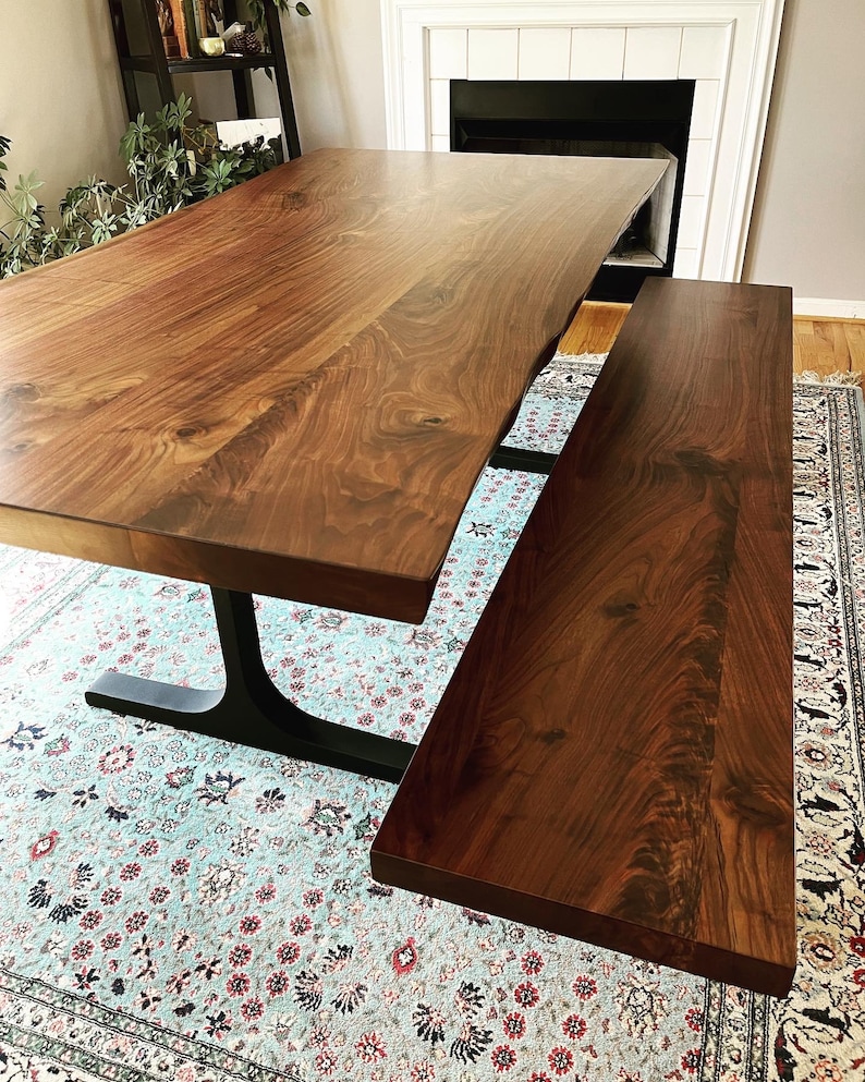May include: A large rectangular dining table and a matching bench made of dark brown wood with a visible natural grain pattern. The table features a live edge side and is supported by a black metal base with a curved, pedestal-style design.