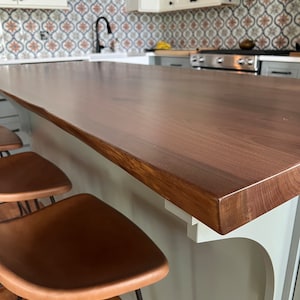 May include: A close-up of a light brown wooden kitchen island countertop with a white bracket. Two brown leather bar stools with black metal legs are in the foreground.