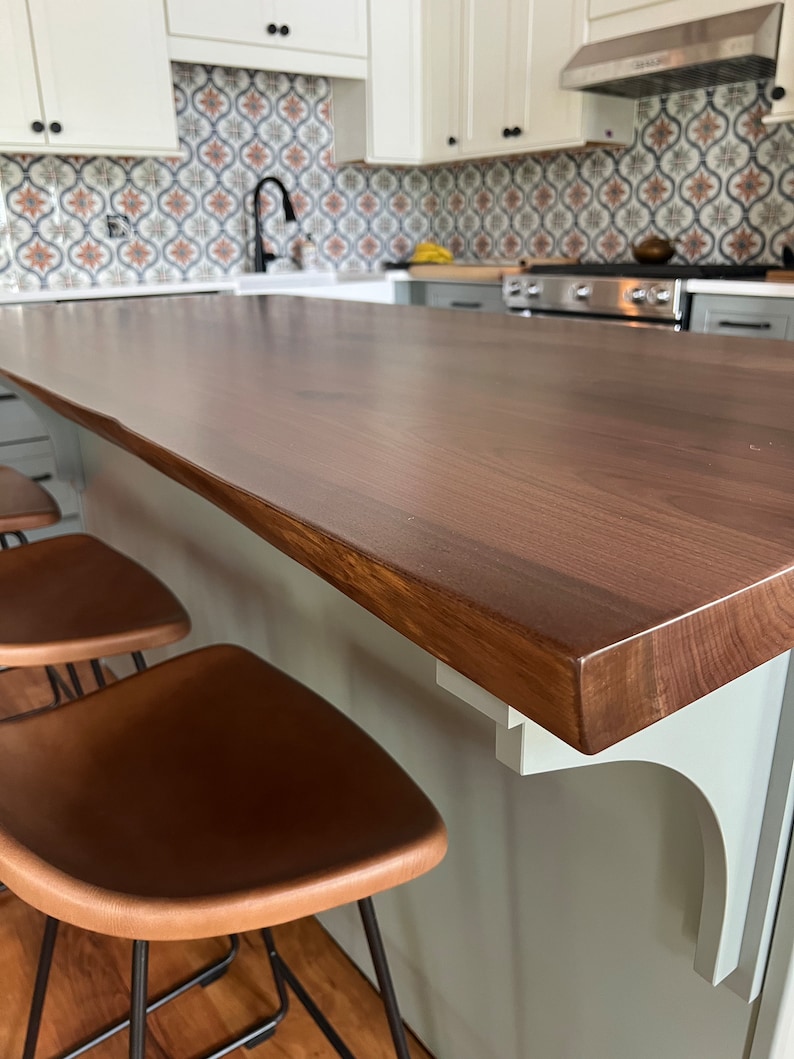 May include: A close-up of a light brown wooden kitchen island countertop with a white bracket. Two brown leather bar stools with black metal legs are in the foreground.