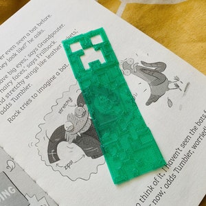 May include: A green Minecraft-themed bookmark featuring a pixelated creeper face. The bookmark is lying on top of a page in a children's book.