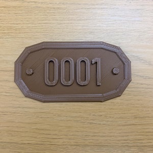 May include: A brown, octagonal sign displaying the number "0001". The sign has a textured surface and a decorative border. Small circular details are either side of the number. The sign is likely for a house or room.