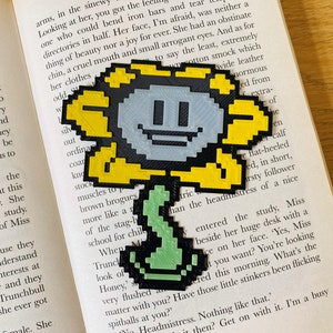 May include: A 3D printed bookmark shaped like a yellow flower with a black outline and a grey face. The flower has a green stem and is smiling. The bookmark is on top of a book with text.