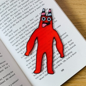 Puede incluir: Un monstruo rojo de dibujos animados con ojos saltones y una amplia sonrisa con dientes. El monstruo está de pie sobre un libro con texto sobre juguetes.