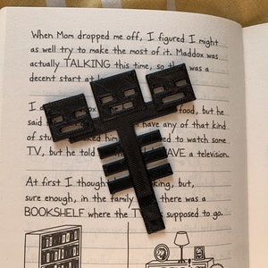 May include: A black 3D printed model of a Minecraft Creeper, a popular video game character. The model is sitting on a page of a book with text about a character named Maddox.