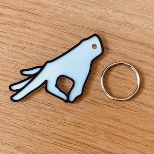 Op de afbeelding: Witte plastic sleutelhanger in de vorm van een hand die de "okay"-gebaren maakt. De sleutelhanger heeft een zwarte omtrek en een zilveren metalen ring.