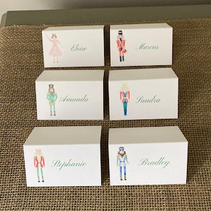 May include: Six white place cards with illustrations of nutcrackers and a ballerina. Each card has a name in green script: Eloise, Marcus, Amanda, Sandra, Stephanie, and Bradley. The cards are arranged on a burlap surface.