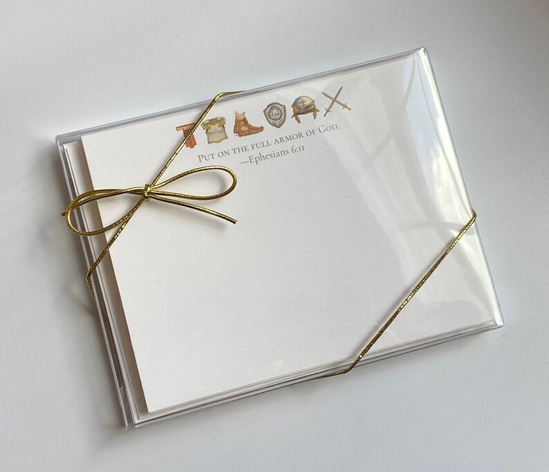Puede incluir: Un juego de tarjetas de notas en blanco con una cinta dorada atada alrededor. Las tarjetas tienen una peque&ntilde;a ilustraci&oacute;n de un conjunto de armaduras y el texto "PUT ON THE FULL ARMOR OF GOD. -Ephesians 6:11"