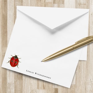 Pode incluir: Conjunto de papelaria branco com uma ilustração de joaninha vermelha, uma caneta dourada e um envelope branco. A papelaria tem o nome "Leslie Witherspoon" impresso em preto. A caneta está em cima da papelaria.