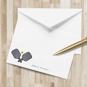 Könnte beinhalten: Weißes Briefpapier mit Pickleball-Motiv. Das Design zeigt zwei gekreuzte graue Pickleball-Schläger und einen gelben Ball. Der Name "John J. Norman" ist unten aufgedruckt. Ein goldener Stift liegt auf dem Briefpapier.