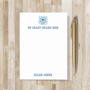 May include: A white notepad with the text "US COAST GUARD MOM" and a Coast Guard emblem in blue. The name "ELLEN JONES" is printed at the bottom. A gold pen rests on a wooden surface.